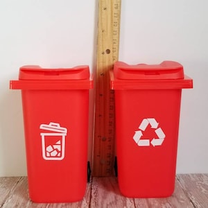 May include: Two red toy trash cans with white lids and wheels. The trash can on the left has a white trash can icon and the trash can on the right has a white recycling symbol.
