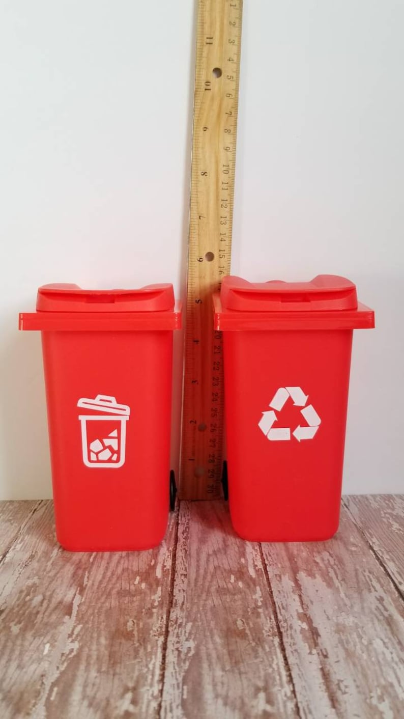 May include: Two red toy trash cans with white lids and wheels. The trash can on the left has a white trash can icon and the trash can on the right has a white recycling symbol.