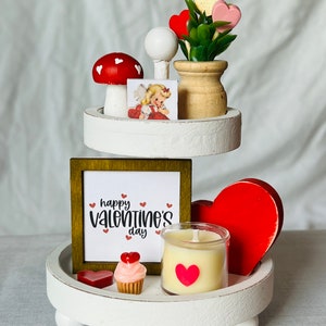 K&ouml;nnte beinhalten: Ein dreistufiger wei&szlig;er Tablett-Set, der f&uuml;r den Valentinstag dekoriert ist. Die oberste Stufe zeigt eine kleine Topfpflanze mit rosa und roten Herzen, einen wei&szlig;en Pilz mit roten Punkten und eine Vintage-Valentinstagskarte. Die mittlere Stufe hat ein gerahmtes Schild mit der Aufschrift "happy valentine's day" mit roten Herzen. Die unterste Stufe hat ein rotes Herz, einen Cupcake mit rosa Zuckerguss und roten Herzen, eine Kerze in einem Glasgef&auml;&szlig; mit einem roten Herzaufkleber und ein kleines rotes Herz.