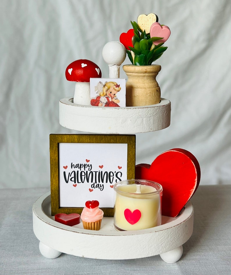 K&ouml;nnte beinhalten: Ein dreistufiger wei&szlig;er Tablett-Set, der f&uuml;r den Valentinstag dekoriert ist. Die oberste Stufe zeigt eine kleine Topfpflanze mit rosa und roten Herzen, einen wei&szlig;en Pilz mit roten Punkten und eine Vintage-Valentinstagskarte. Die mittlere Stufe hat ein gerahmtes Schild mit der Aufschrift "happy valentine's day" mit roten Herzen. Die unterste Stufe hat ein rotes Herz, einen Cupcake mit rosa Zuckerguss und roten Herzen, eine Kerze in einem Glasgef&auml;&szlig; mit einem roten Herzaufkleber und ein kleines rotes Herz.