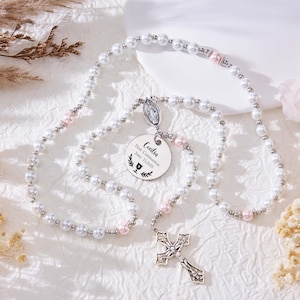 May include: A pearl rosary with a silver cross pendant and a round tag engraved with the name "Cecilia" and "First Communion". The rosary features white and pink beads, with a decorative silver pendant. The image is set against a white background with floral accents.