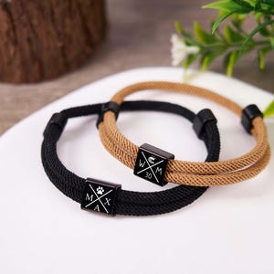 Op de afbeelding: Twee touwarmbanden, één zwart en één bruin, met zwarte vierkante sluitingen. De zwarte armbandsluiting heeft een pootafdruk en de letters MAX. De bruine armbandsluiting heeft een logo en het nummer 30.