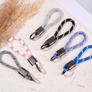 Puede incluir: Una colección de llaveros con bucles de cuerda trenzada en varios colores: gris, negro, morado, azul y un patrón blanco y coral. Cada llavero tiene un cierre de metal y un conector metálico rectangular.