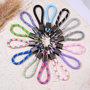 Puede incluir: Una colección de llaveros coloridos con lazos de cuerda trenzada. Los llaveros presentan una variedad de colores, incluyendo azul, verde, rosa, morado y negro. Cada llavero tiene un cierre de metal y un lazo para facilitar la fijación.