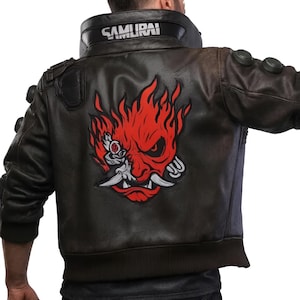 Könnte beinhalten: Eine dunkelbraune Lederjacke mit einer großen rot-schwarzen Grafik einer stilisierten Oni-Maske auf dem Rücken. Der hohe Kragen zeigt das Wort SAMURAI in weißen Buchstaben. Die Jacke hat dunkle, kreisförmige Akzente an Schultern und Ärmeln.