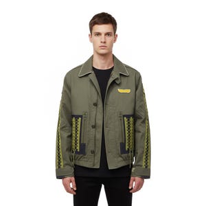Puede incluir: Chaqueta verde oliva con cierre de botones, ribete blanco en el cuello y parche amarillo en el pecho. Las mangas y los bolsillos presentan patrones decorativos en negro y amarillo. La chaqueta se lleva sobre una camiseta negra.