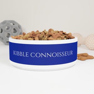 Puede incluir: Un cuenco blanco de cerámica para mascotas con una banda azul vibrante con el texto "KIBBLE CONNOISSEUR" en blanco. El cuenco está lleno de comida para perros. Un juguete gris y un juguete en forma de hueso rosa claro están en el fondo.