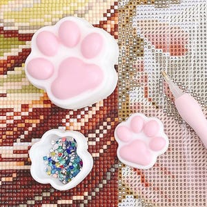May include: Three white and pink paw-shaped trays for diamond painting. One tray is filled with colourful diamond painting gems. A pink stylus is also visible, along with a diamond painting canvas in the background.