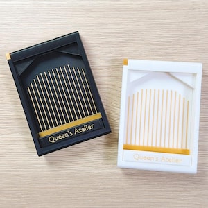 May include: Two rectangular, decorative boxes, one black and one white, with gold accents. Each box features vertical gold lines and the text "Queen's Atelier" on a gold band.