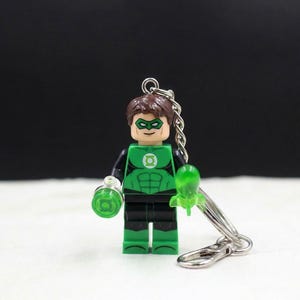 May include: A Green Lantern Lego figure keychain. The figure is primarily green and black, with a brown hair piece and a Green Lantern logo on the chest. It holds a green lantern and a green energy construct. The keychain has a silver chain and clasp.