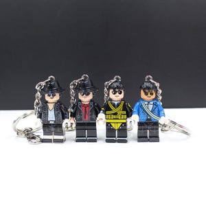 May include: Four Lego-style keychains featuring miniature figures in various outfits, including a black jacket with silver studs, a red shirt and black jacket, a yellow and black military-style jacket, and a blue jacket with white accents. Each figure has a black hat and sunglasses.