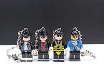 May include: Four Lego-style keychains featuring miniature figures in various outfits, including a black jacket with silver studs, a red shirt and black jacket, a yellow and black military-style jacket, and a blue jacket with white accents. Each figure has a black hat and sunglasses.