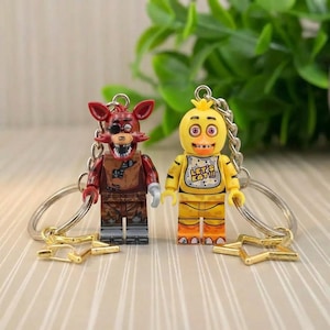May include: Two keychains featuring miniature toy figures. One is a red fox-like character with a pirate theme, and the other is a yellow chicken character with the text "LET'S EAT!!!" on its bib. Both have silver key rings.