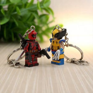 May include: Two keychains featuring Lego-style figures of Deadpool and Wolverine. Deadpool is in a red and black suit, holding a gun. Wolverine is in a yellow and blue suit, with claws extended. Both figures are attached to silver keyrings.