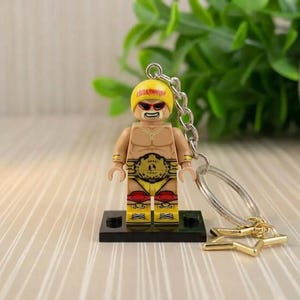 May include: A yellow and red wrestler figurine keychain. The figurine has a yellow hat with the word "HULKAMANIA" printed on it, red sunglasses, and a championship belt. The keychain has a gold star charm.
