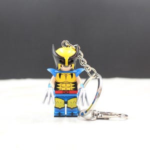 May include: A Wolverine-themed keychain featuring a minifigure with a yellow and black mask, blue and yellow costume, and silver claws. The keychain has a silver chain and clasp, perfect for attaching to keys or a backpack.