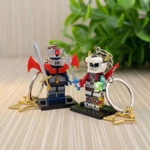 May include: Two robot-themed keychains. One robot is red, blue, and gray, holding a sword. The other robot is red, white, and yellow, also holding a sword. Both keychains have gold-colored star-shaped accents.