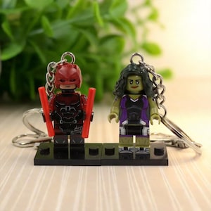 May include: Two superhero-themed keychains. One features a red and black character with red batons, the other a green-skinned character in purple and black attire. Both figures are attached to keychains and mounted on black bases.