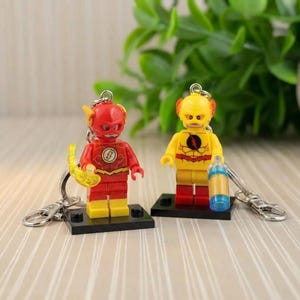 May include: Two Lego-style keychains featuring superhero figures. One is red and yellow, holding a yellow lightning bolt. The other is yellow and red, holding a blue and clear cylinder. Both figures stand on black bases with silver keychains.