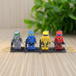 May include: Four miniature action figure keychains in silver, blue, yellow, and red. Each figure is holding a black rifle and attached to a keychain with a star-shaped charm. The figures are standing on black bases.