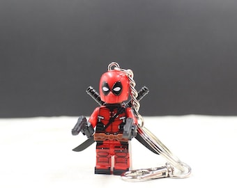 May include: A red and black Deadpool Lego figure keychain. The figure has two black swords on its back, two black guns, and a silver keychain attachment. The figure is standing on a white surface with a dark gray background.