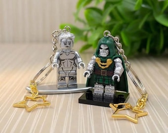 May include: Two superhero-themed keychains. One keychain features a silver-colored figure with a star-shaped charm. The other keychain has a figure with a green hood and a gold star-shaped charm. Both figures are on a base.