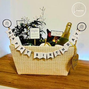 Könnte beinhalten: Ein beiger Weidenkorb gefüllt mit einer Flasche Sekt, einer kleinen Pflanze und anderen Geschenken. Der Korb ist mit einem "Happy Birthday" Banner und kleinen Schildern mit Text dekoriert. Der Korb steht auf einer Holzoberfläche.