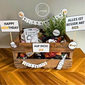 May include: A wooden crate filled with beer bottles, snacks, and greenery, decorated with signs that say "Happy Beerthday", "Alles ist besser mit Bier", and "Auf Dich!". The crate has a banner that says "Biergarten" and "Prost".