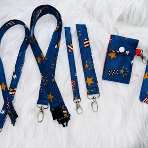 May include: A set of patriotic-themed accessories, including two lanyards, two wristlets, a cardholder, and a phone case. The items are navy blue with a pattern of gold stars, white stars, and red and white stripes.