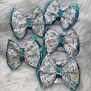 May include: Five hair bows featuring a white fabric base with a colorful mermaid pattern, accented by numerous multicolored rhinestones. Each bow has a metallic teal border and a central knot embellished with a cluster of sparkling crystals.