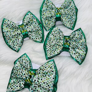 May include: Four hair bows featuring a light green base covered in a dense pattern of sparkling clear, dark green, and light green rhinestones. Each bow has a metallic green center band and a metallic green border along the outer edges.