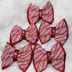 May include: Five hair bows featuring a red and white diagonal striped pattern. Each bow is embellished with scattered clear rhinestones and a central red fabric knot adorned with a cluster of sparkling crystals at the center of the bow.