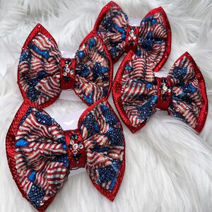 May include: Five red, white, and blue hair bows with an American flag design. Each bow features a red glitter border and a center adorned with rhinestones. The bows are arranged on a white, fluffy surface.