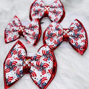 May include: Four patriotic hair bows. Each bow features a white background with red and blue flag-themed patterns. The bows have a red border and a sparkling center embellishment. Perfect for celebrating Independence Day or other patriotic events.