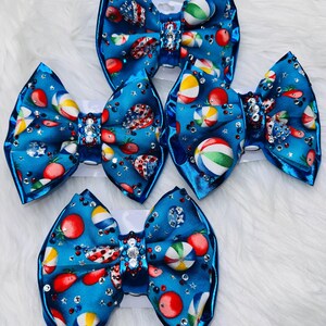 May include: Five blue hair bows with a summer theme. Each bow features a pattern of strawberries, beach balls, and watermelon slices. The bows are embellished with rhinestones and have a shiny, satin-like finish.