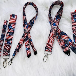 May include: A collection of patriotic lanyards and a phone case, all featuring a repeating pattern of the American flag. The lanyards have metal clasps and black adjustable buckles. The items are displayed on a fluffy white surface.