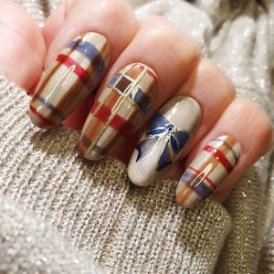Puede incluir: Una mano con uñas largas y ovaladas. Las uñas presentan un diseño a cuadros en tonos marrones, rojos y azules. Una uña tiene un diseño de lazo azul. Las uñas están pintadas con un acabado brillante.