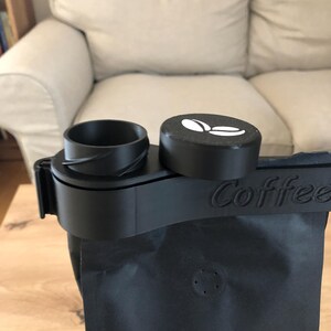 Puede incluir: Dispensador de bolsas de caf&eacute; negro con tapa negra y logotipo de grano de caf&eacute; blanco. El dispensador tiene una correa negra con la palabra "Coffee" impresa.