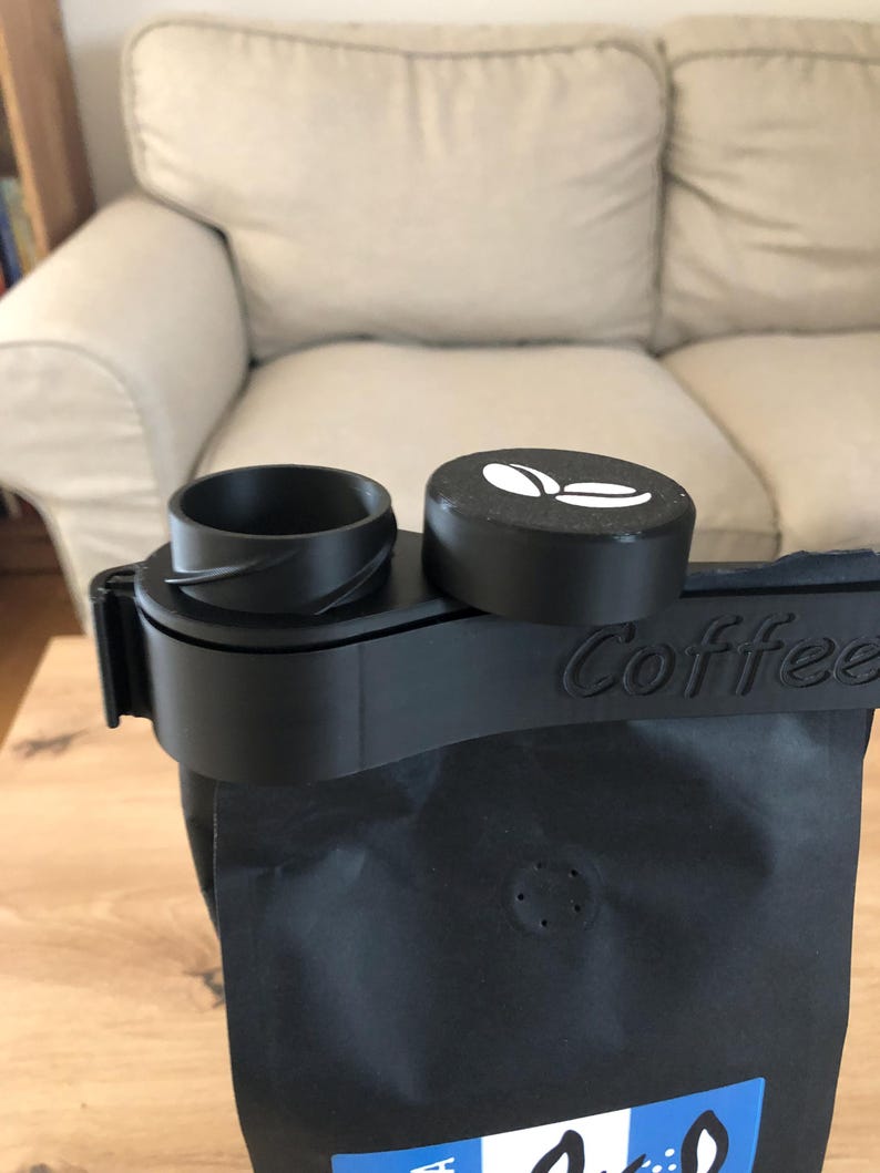 Puede incluir: Dispensador de bolsas de caf&eacute; negro con tapa negra y logotipo de grano de caf&eacute; blanco. El dispensador tiene una correa negra con la palabra "Coffee" impresa.