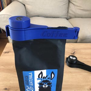 Puede incluir: Clip azul para bolsa de caf&eacute; con la palabra "Coffee" impresa en &eacute;l. El clip est&aacute; unido a una bolsa negra de granos de caf&eacute; con una ilustraci&oacute;n de llama y el texto "100% Arabica Guatemala".