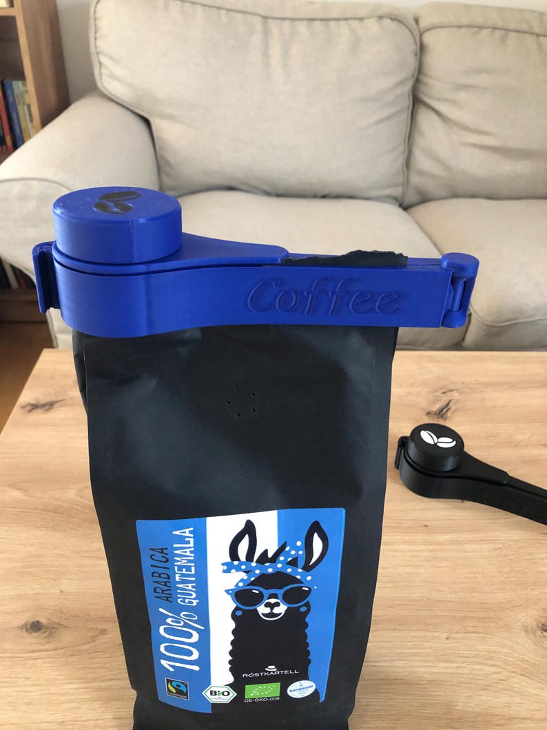 Puede incluir: Clip azul para bolsa de caf&eacute; con la palabra "Coffee" impresa en &eacute;l. El clip est&aacute; unido a una bolsa negra de granos de caf&eacute; con una ilustraci&oacute;n de llama y el texto "100% Arabica Guatemala".