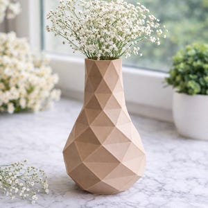 Puede incluir: Un jarrón beige geométrico impreso en 3D lleno de flores blancas. El jarrón tiene un patrón de diamantes y está sobre una superficie de mármol. El texto "Geometric 3D-Printed Vase" está en la parte superior de la imagen.