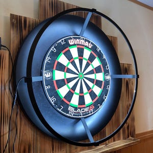 Puede incluir: Un tablero de dardos negro con el logotipo de Winmau Blade 5 y un marco de metal negro con una luz de halo. El tablero de dardos está montado en una pared de madera.