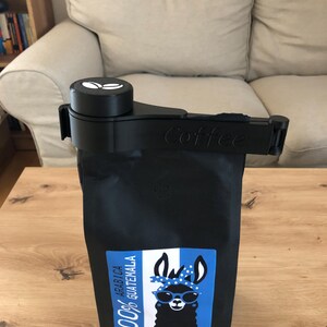 Puede incluir: Porta bolsas de caf&eacute; negro con un logotipo de grano de caf&eacute; blanco y la palabra "Coffee" impresa en el lateral. La bolsa est&aacute; llena de granos de caf&eacute; 100% Ar&aacute;bica Guatemala y presenta una llama de dibujos animados con gafas de sol en la etiqueta.
