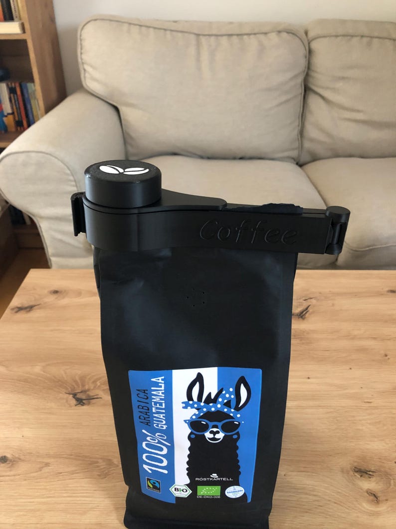 Puede incluir: Porta bolsas de caf&eacute; negro con un logotipo de grano de caf&eacute; blanco y la palabra "Coffee" impresa en el lateral. La bolsa est&aacute; llena de granos de caf&eacute; 100% Ar&aacute;bica Guatemala y presenta una llama de dibujos animados con gafas de sol en la etiqueta.
