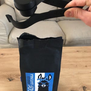 Puede incluir: Una bolsa de caf&eacute; negra con una llama con gafas de sol en la parte delantera. La bolsa est&aacute; etiquetada "100% Arabica Guatemala".
