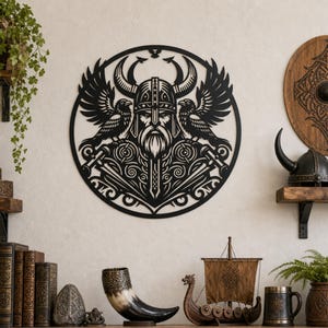 Peut inclure: Une décoration murale circulaire en métal noir présentant une découpe détaillée d'un guerrier barbu portant un casque à cornes, flanqué de deux corbeaux aux ailes déployées, fixée sur un mur clair avec divers objets décoratifs en dessous.