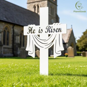 May include: White wooden cross with the text "He is Risen" and a draped fabric design. The cross is placed in a grassy area in front of a church building. A religious item.