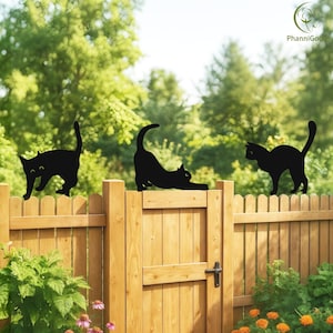 May include: Three black cat silhouettes are mounted on a wooden fence. One cat is walking, another is stretching, and the third is standing. The fence is made of light brown wood, with green foliage and a clear sky in the background.