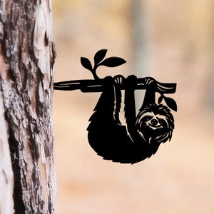 Puede incluir: Una silueta de metal negro de un perezoso colgado de una rama de árbol horizontal. El perezoso se muestra con los brazos envueltos alrededor de la rama, con texturas de pelaje detalladas y una cara sonriente. Dos hojas pequeñas en la rama.